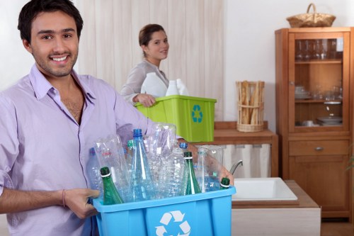 A man with dark hair, a beard, and a friendly expression, wearing a light purple shirt, is holding a blue recycling bin filled with empty glass bottles and jars, placed in a kitchen with a wooden cupboard and a white sink. Behind him, a woman with brown hair tied back, dressed in a grey top, stands near a green recycling bin with a recycling symbol, positioned on a wooden countertop. The kitchen has a neutral colour palette with light curtains and a wooden cabinet with glass doors. The scene captures a moment of household rubbish sorting, consistent with domestic rubbish removal activities in Sydenham, London, as part of professional house clearance carried out by Sydenham House Clearance.